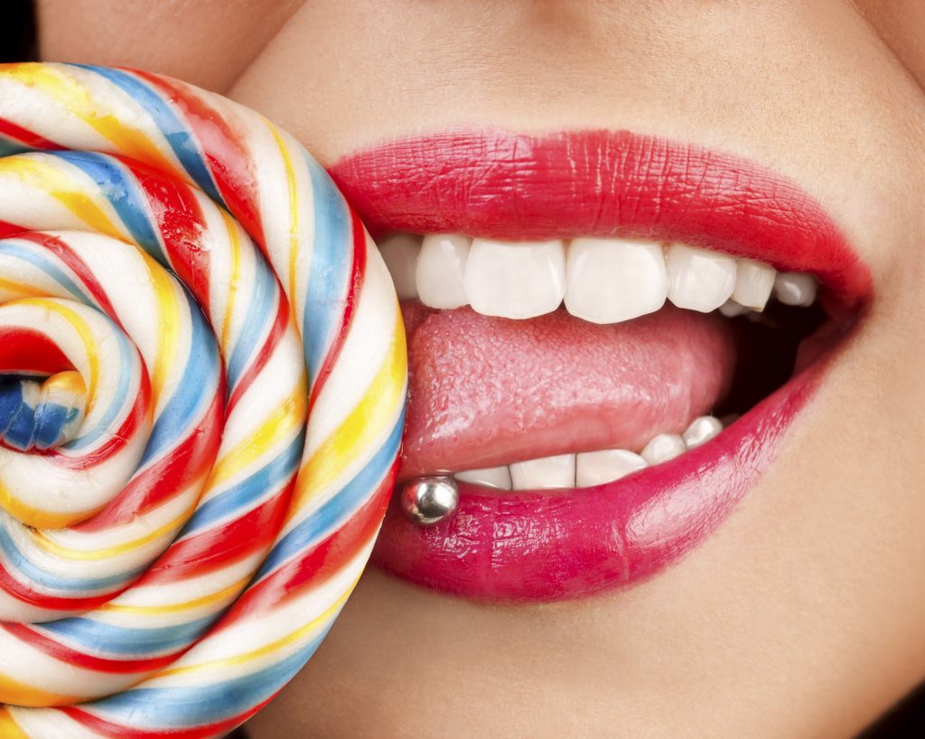 Beautiful girl licking handmade lollipop with pierced tongue, having different color lipstick on lips.
